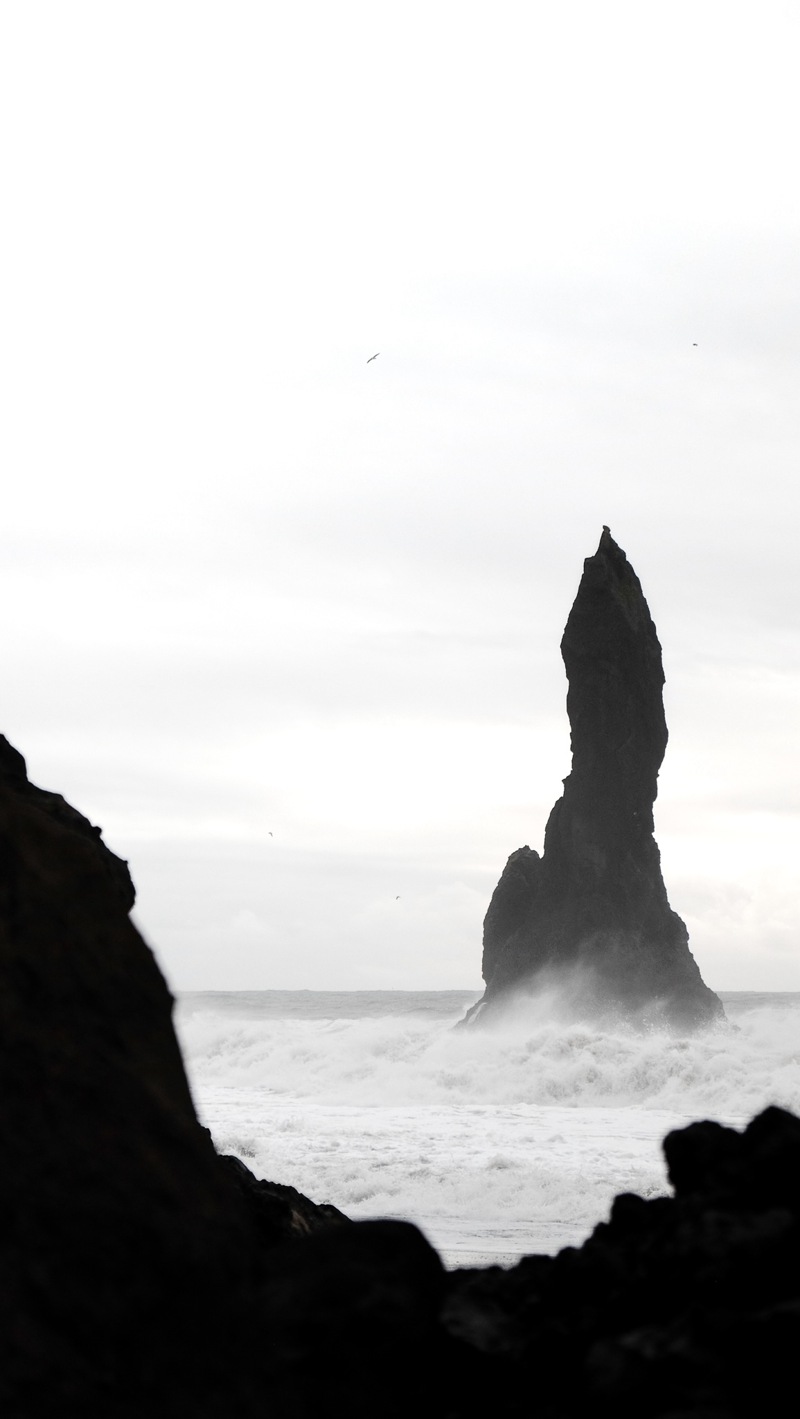 Phyallic looking black rock is surrounded by energetic white foamy sea waves and a white overcast sky, which is framed by black rocks, birds flying in the wind against the white sky