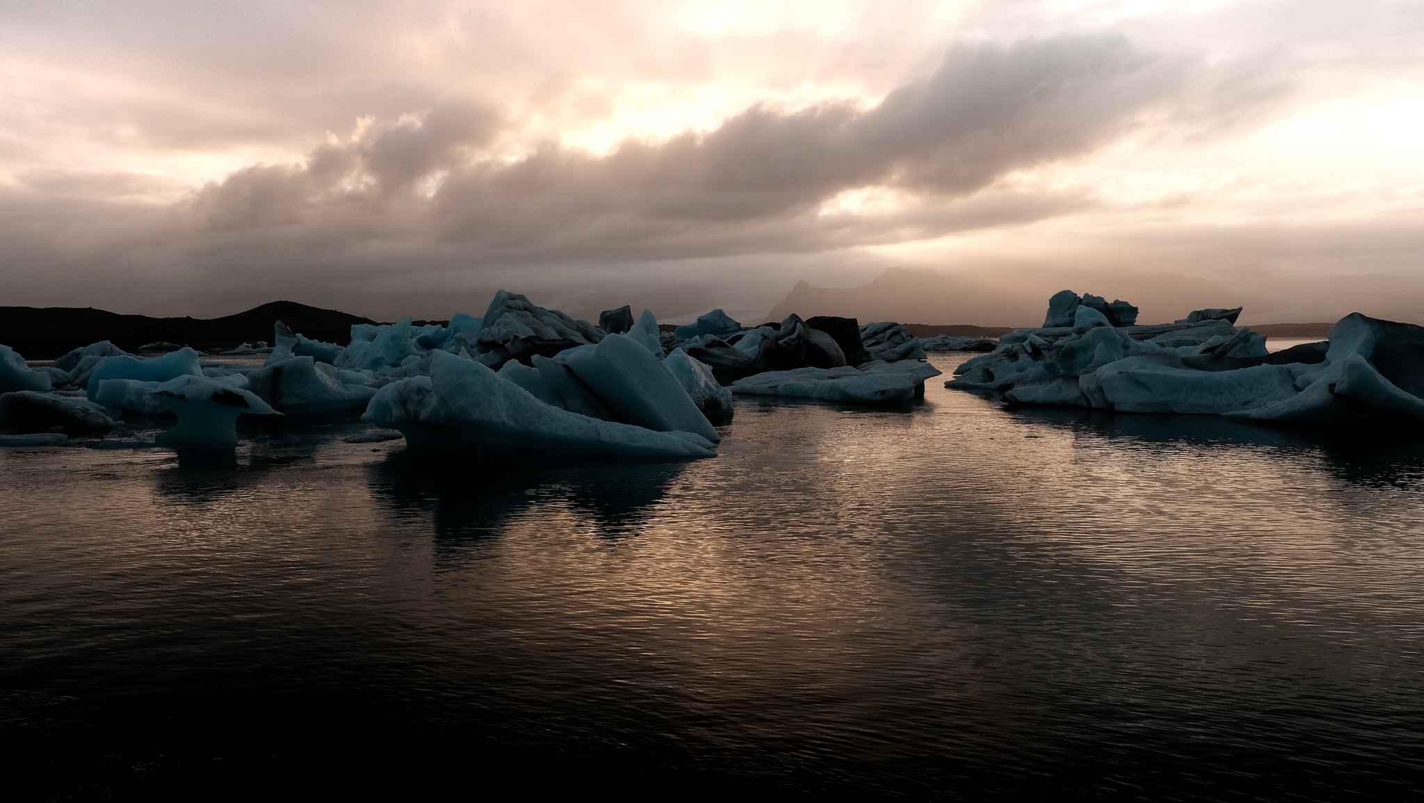 Sun sets behind a glaciel lake reflecting on the water, with large blue ice chunks float