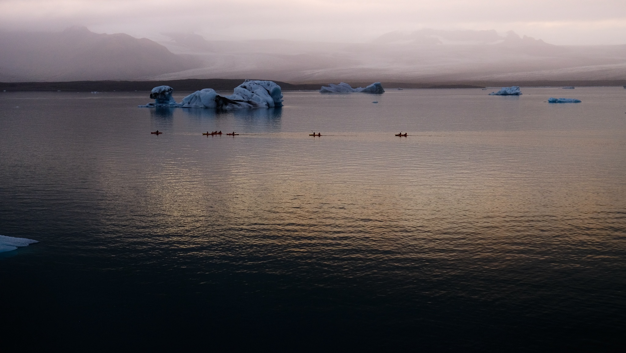 A group of kayaking tourists moving in the distance after the sun has set, illuminated by the evening sky