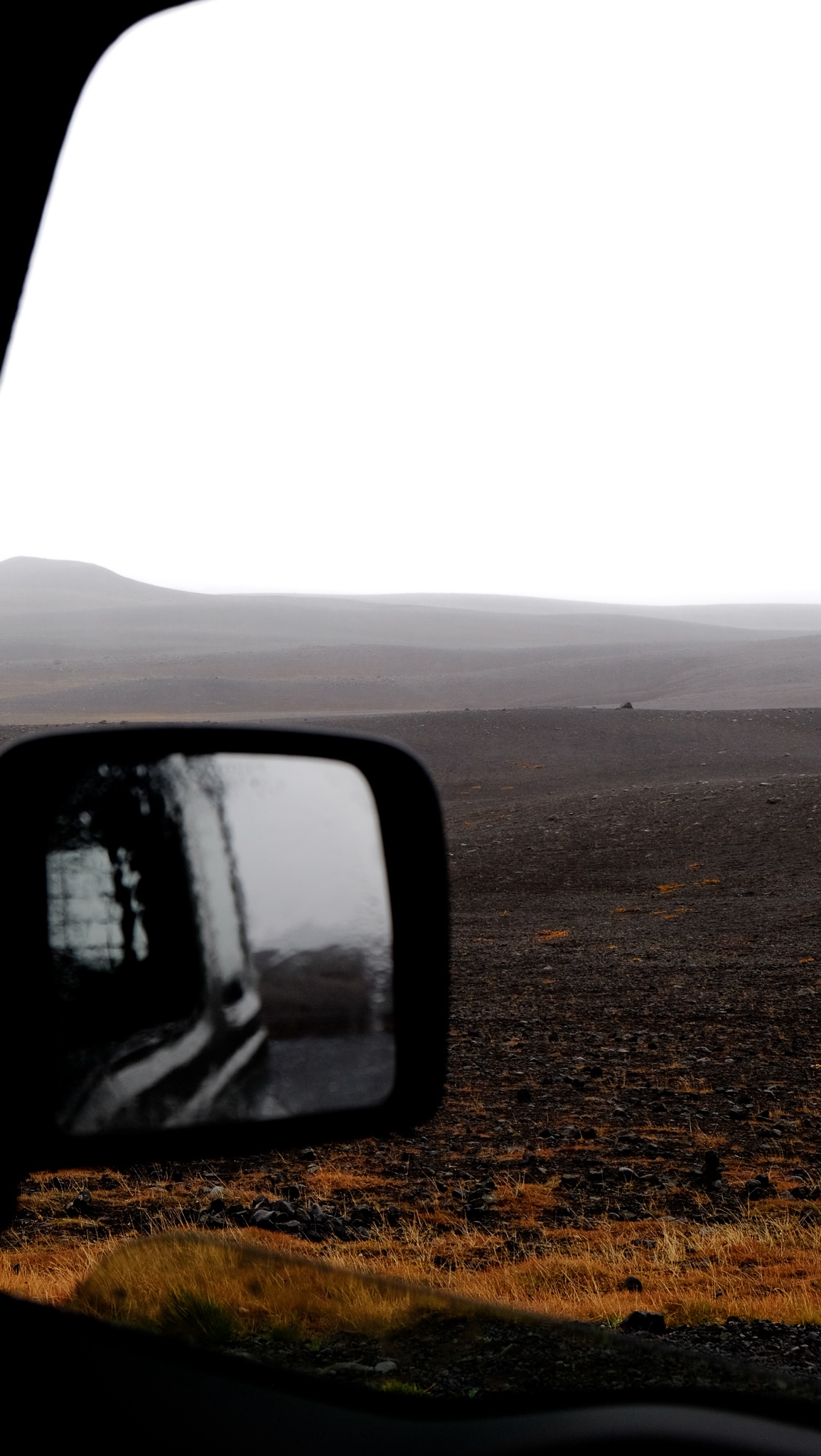 Barren mountain pass battered by wind and rain fades to a white sky, water droplets cover the sideview mirror of the car from which the photo is taken