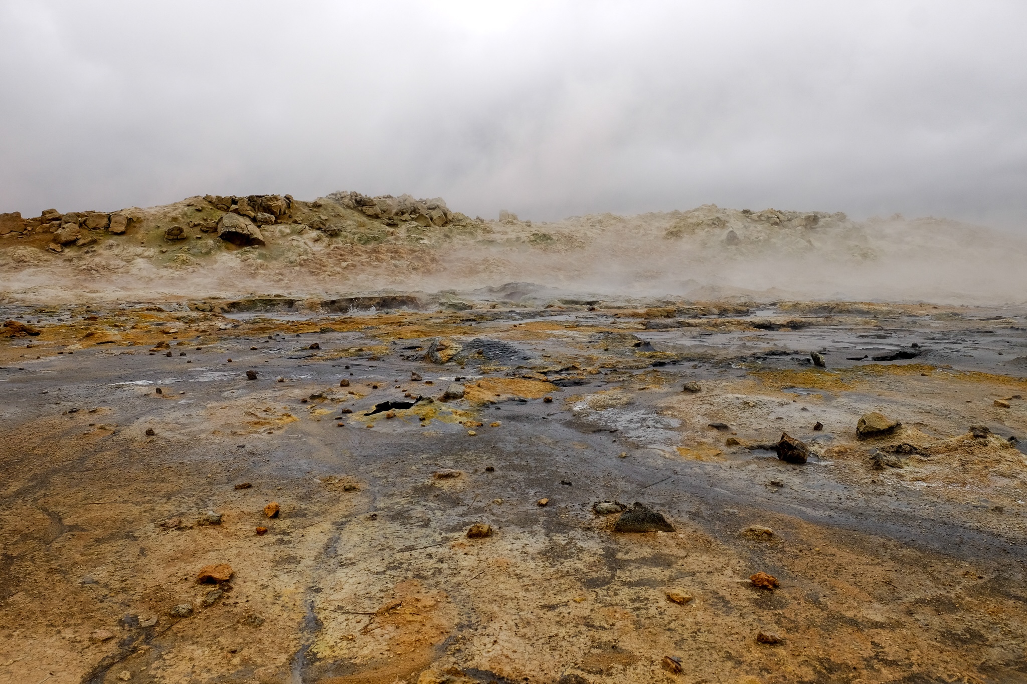 Orangy mineral deposits contrast black deposits with mist floating over the geothermal area