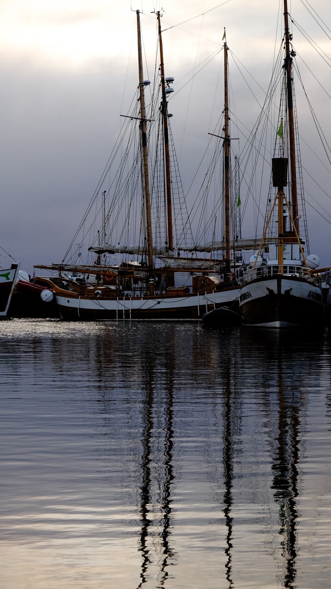 Mast and rope of wooden sailing boats reflect in the water in a harbour