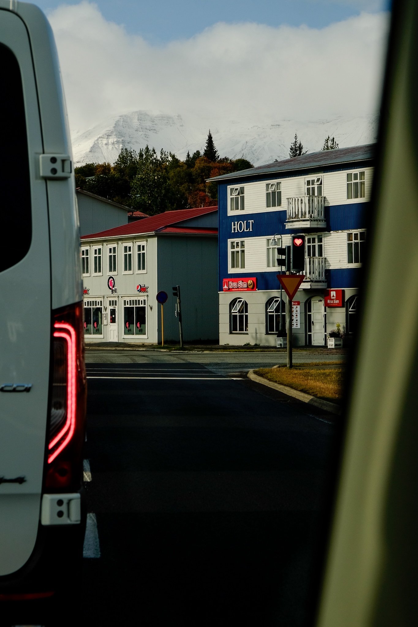A traffic light shining red is in the shape of a heart with buildings and snow capped mountain peaks in the background