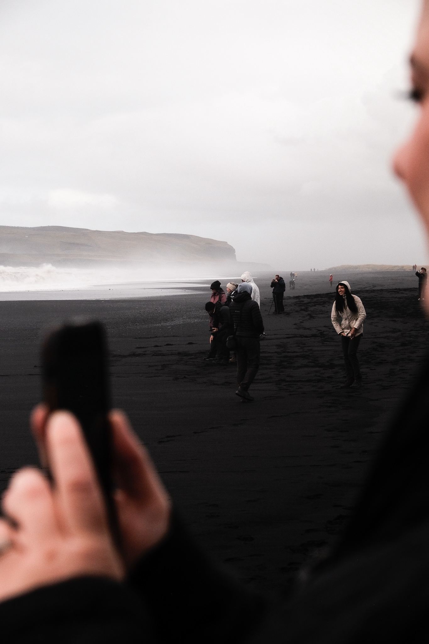 People smiling at each other scattered across the black beach as they fade out into the distance the further out they are, in the foreground, Jenna's taking a timelapse of the sea