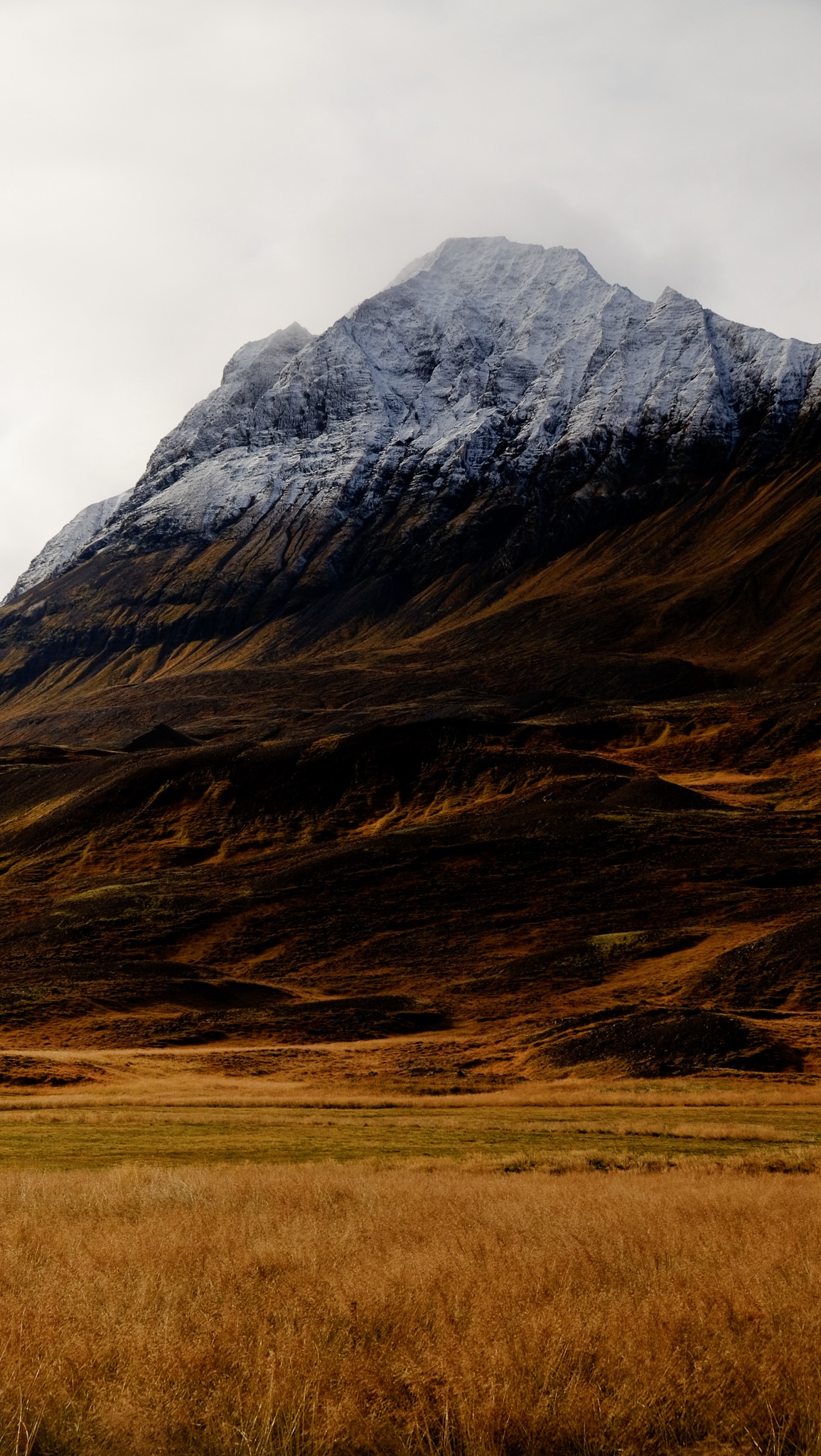 A gradient of colour from pale yellow grasses, to brown mountain side into snow capped peaks and the clouds overhead