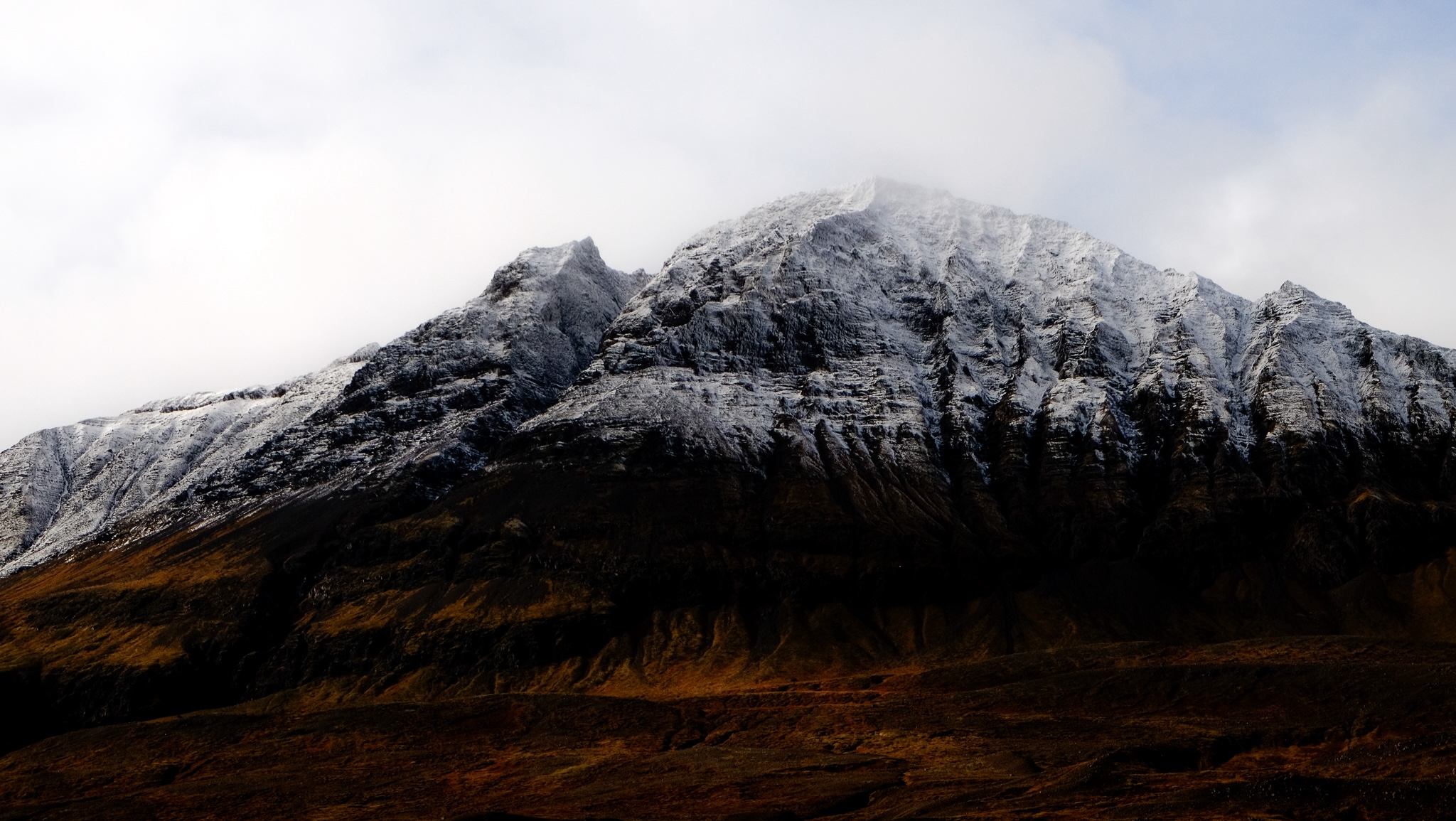 A gradient of colour from a deep brown mountain side into snow capped peaks and the clouds overhead