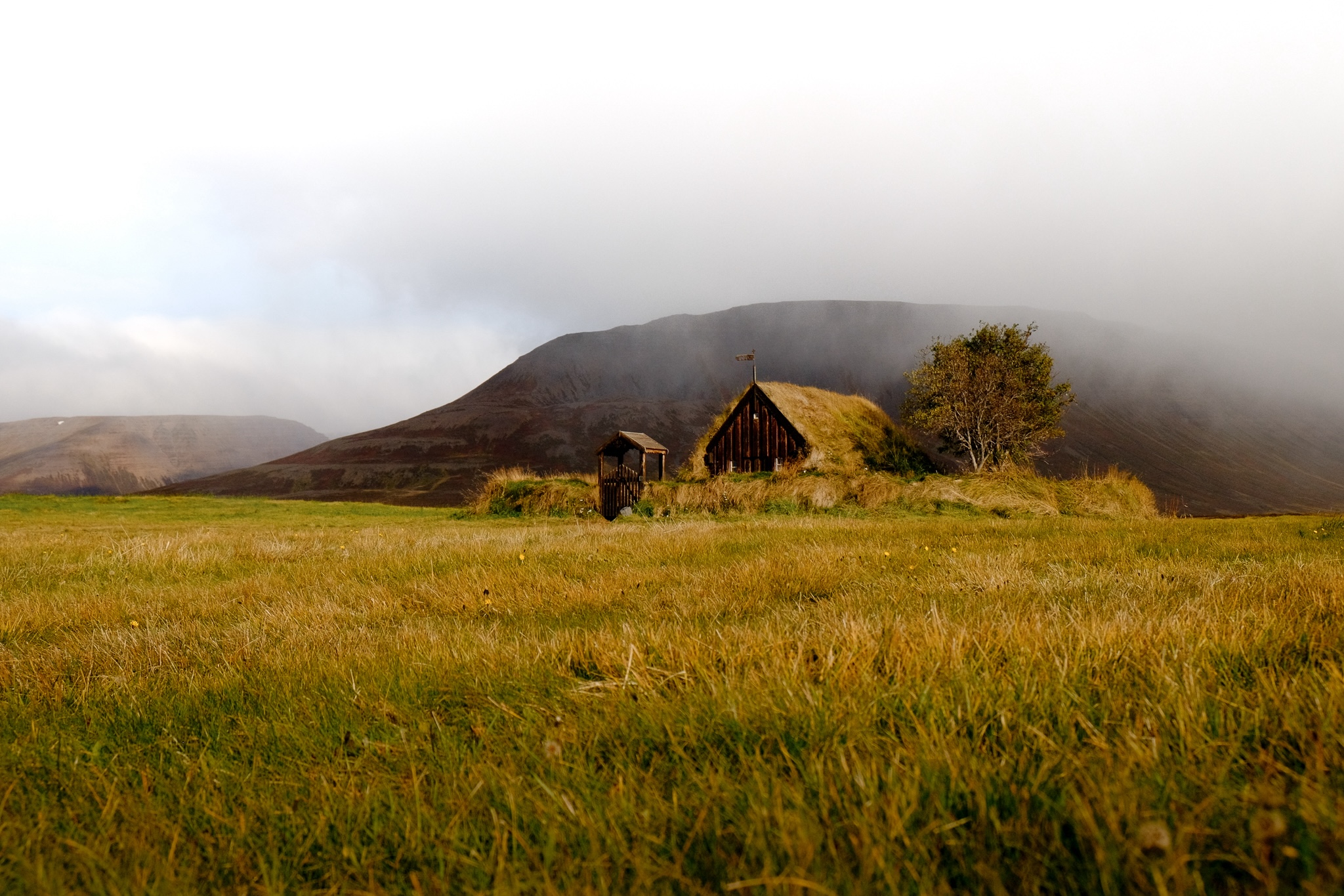 An old church with a grass roof and circular enclosure and a tree, in the distance from a grass field with a mountain in the background with misty clouds rolling in
