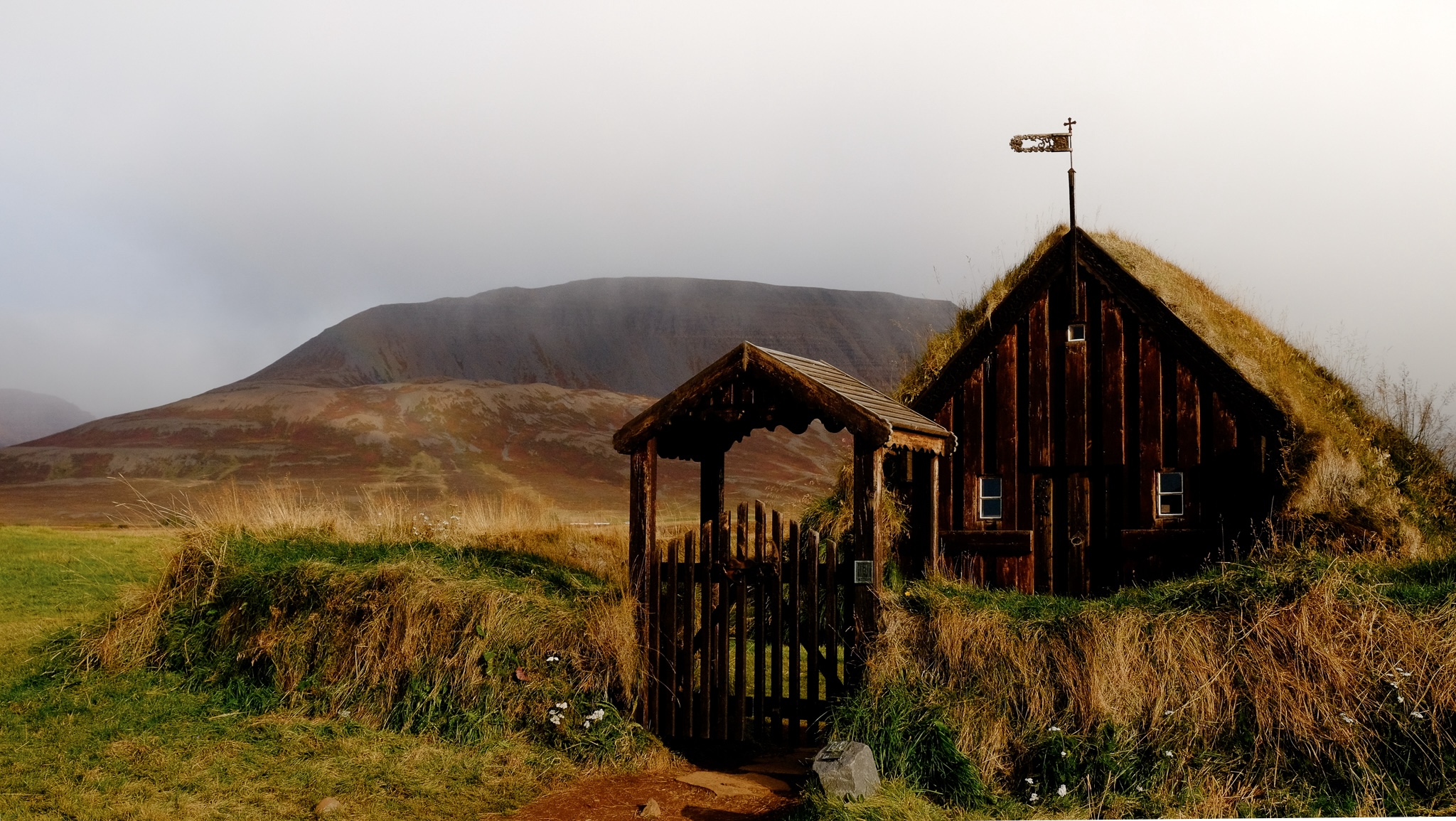 A wooden gate leads to an old brown wooden church covered in grass inside a circular enclosure