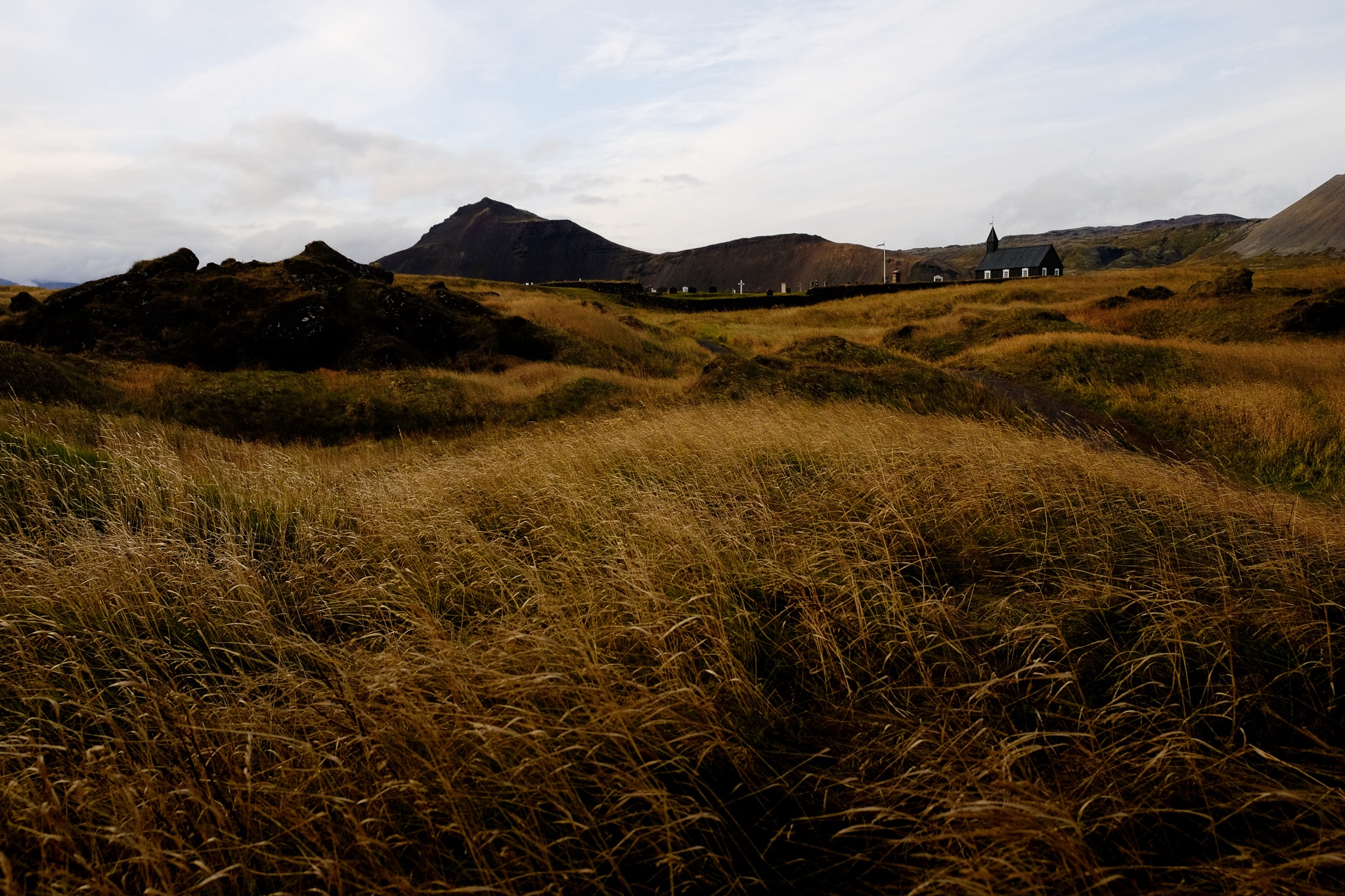 A long grass covered landscape of rolling hills leads to a black church with striking a constrast of white window frames