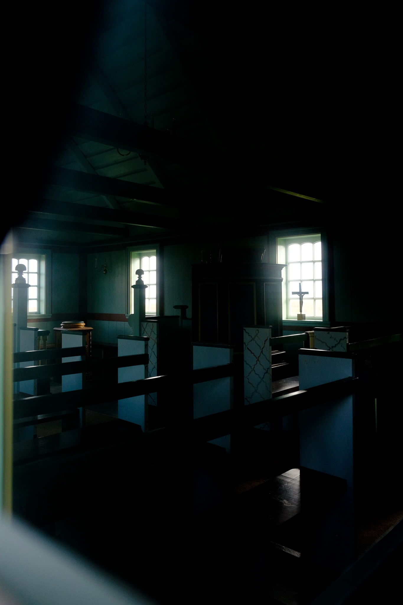 In the window of a church are wooden pews with a white endings, the interior has a cool blue tinge, and an open roof with exposed beams