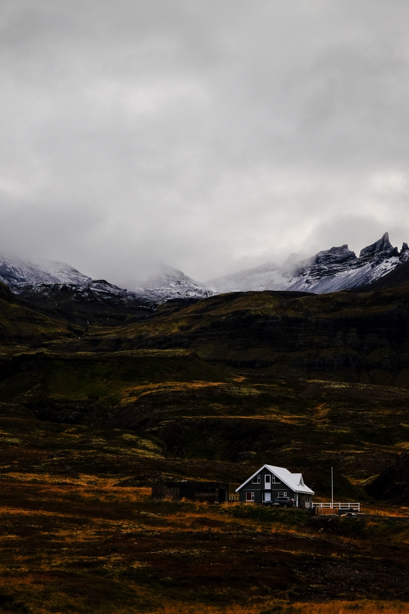 A dark house with a white roof and white trim stands small on a mountainside as the mountain rises into the sky ending with snow capped peaks fading into the clouds