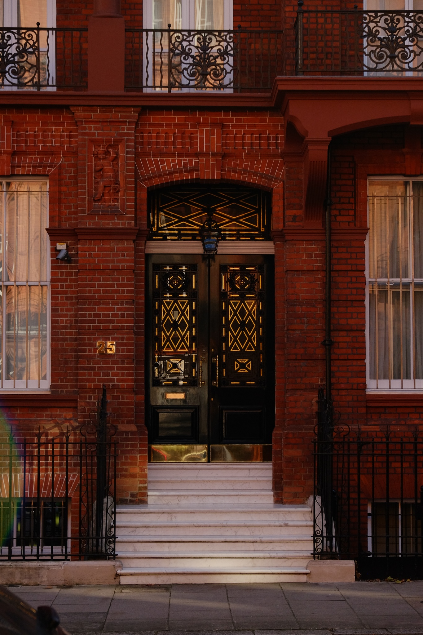 Bright red brick building front door entrance head on, number 25. The door is black with gold diamond patterns over the glass. The stairs from the concrete sidewalk are covered in white marble.