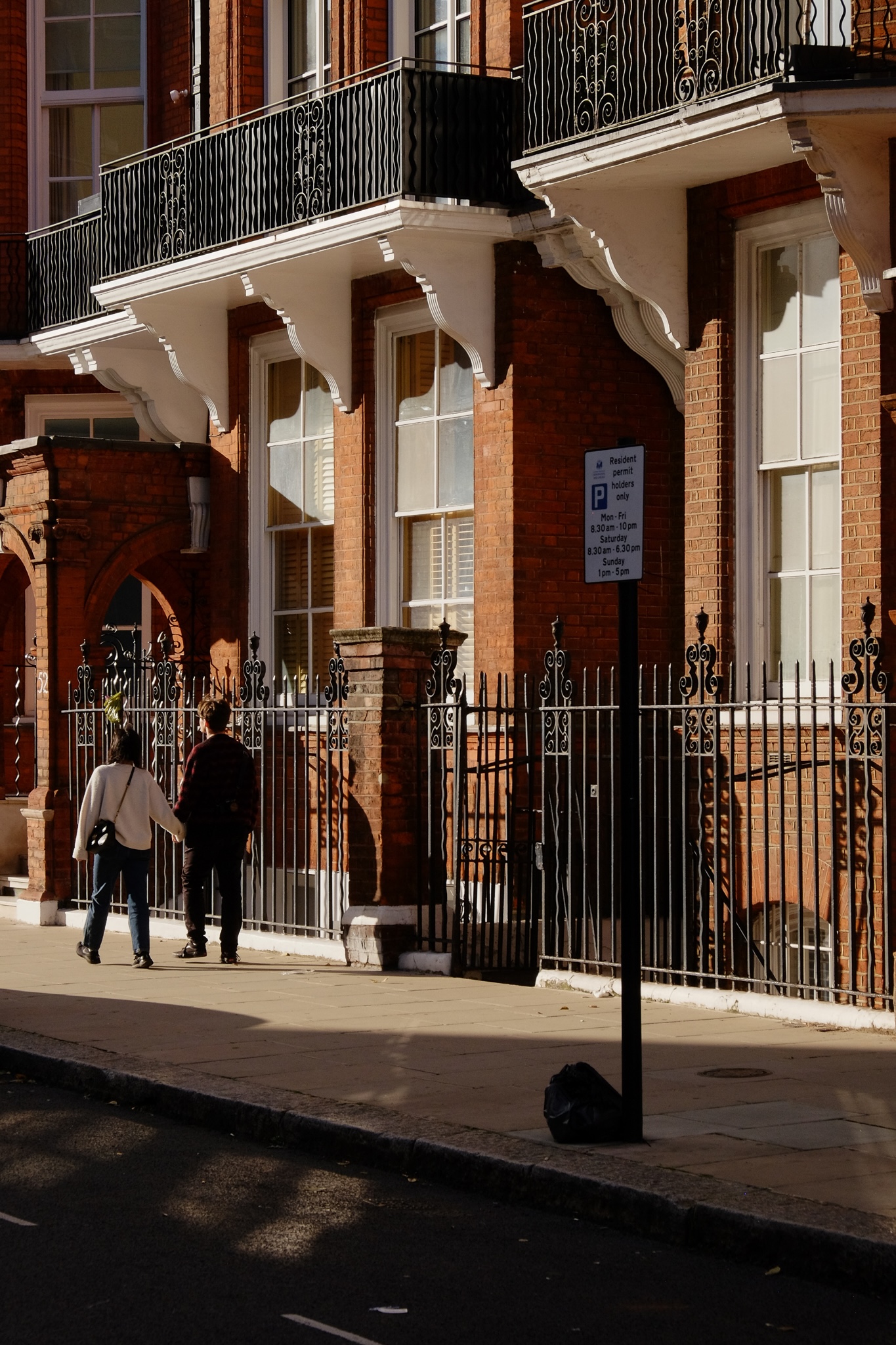 A man and women hold hands walking down the sidewalk with a hard shadown shining on them from the sun, the shade is dark, and there is a red brick building with white trim and black steel work in the background.