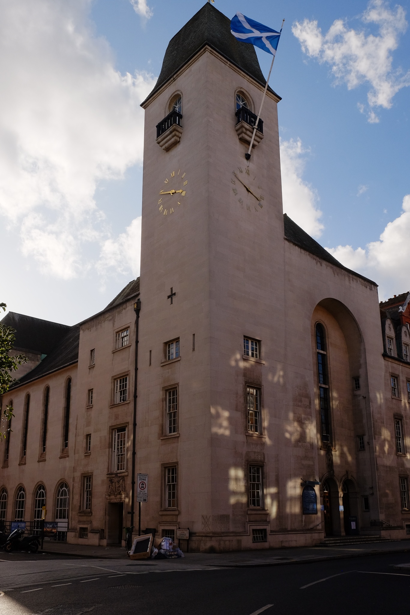 A white concrete Scottish church town stands across the road with the Scottish flag waving and a gold roman numeral clock that is built onto the concrete of the building shines.