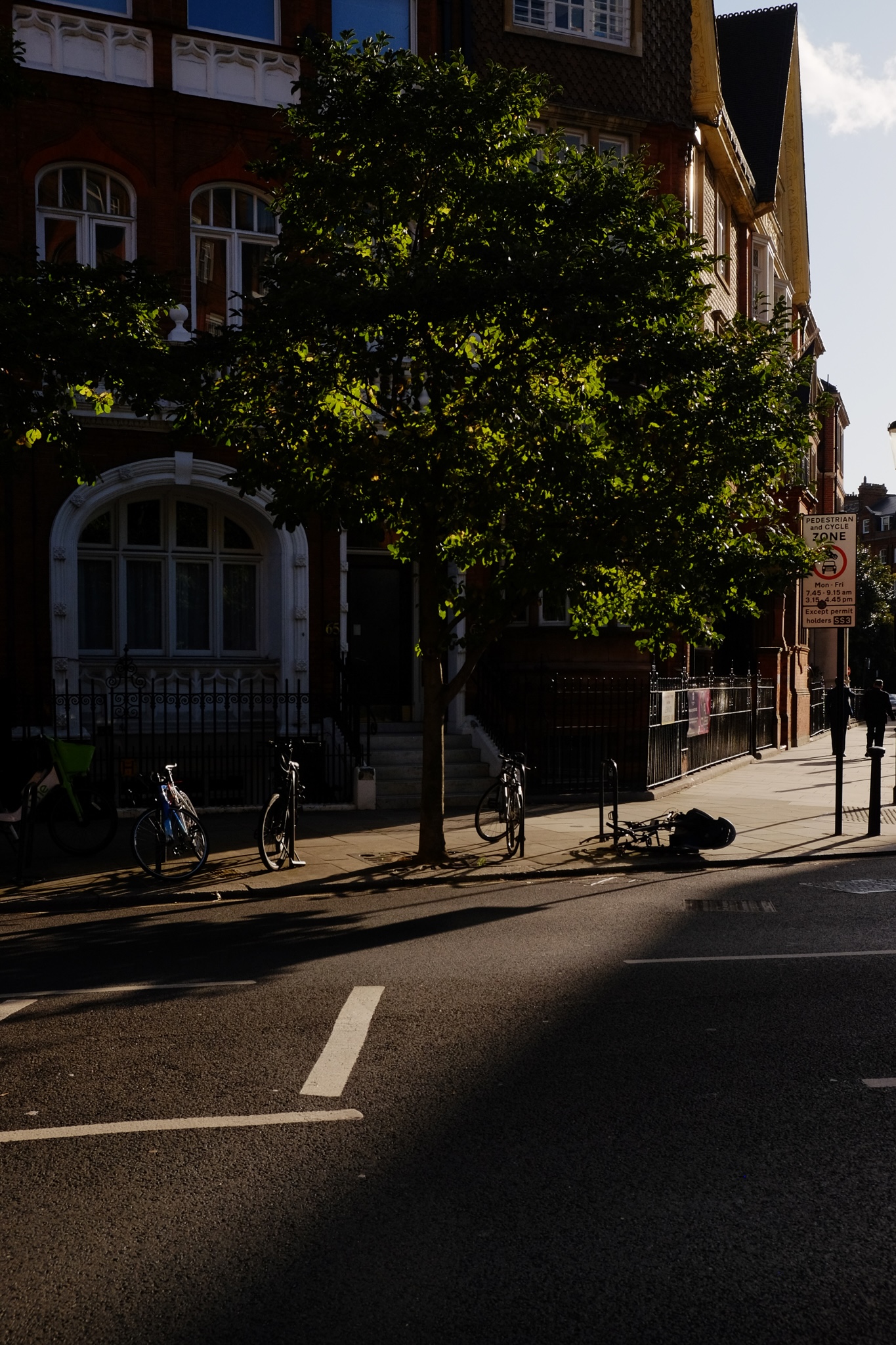 A hard afternoon shadown cuts across a road and sidewalk casting long shadows as two people walk side by side along the sidewalk in the warming sun.
