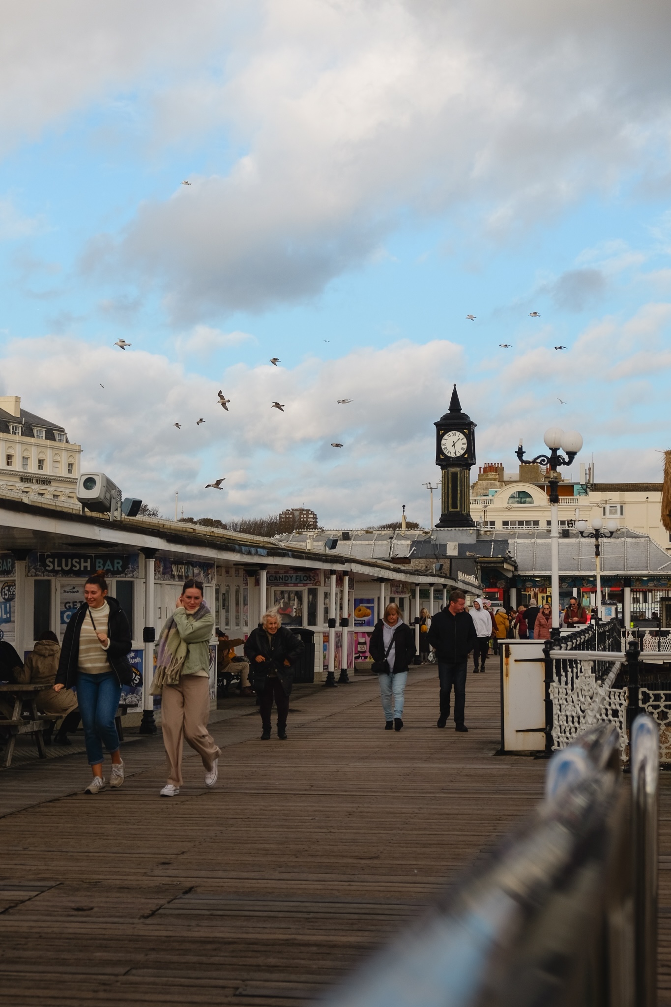 People walking onto the pier are happy with birds flying overhead and an analog clock stands in the distance