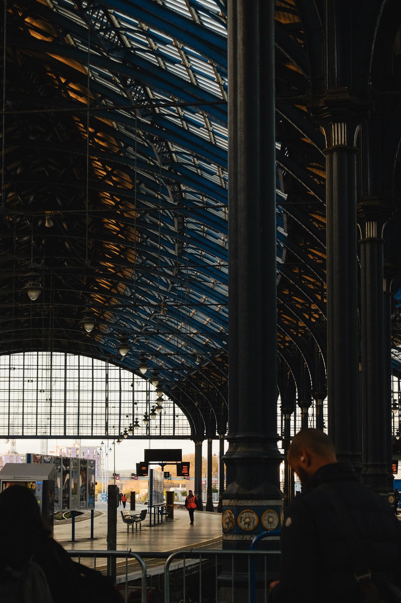 An empty train platform with officials wearing bright orange vests, the ornate roof of the station creates pleasing patterns, two people in the foreground are waiting for their train to arrive